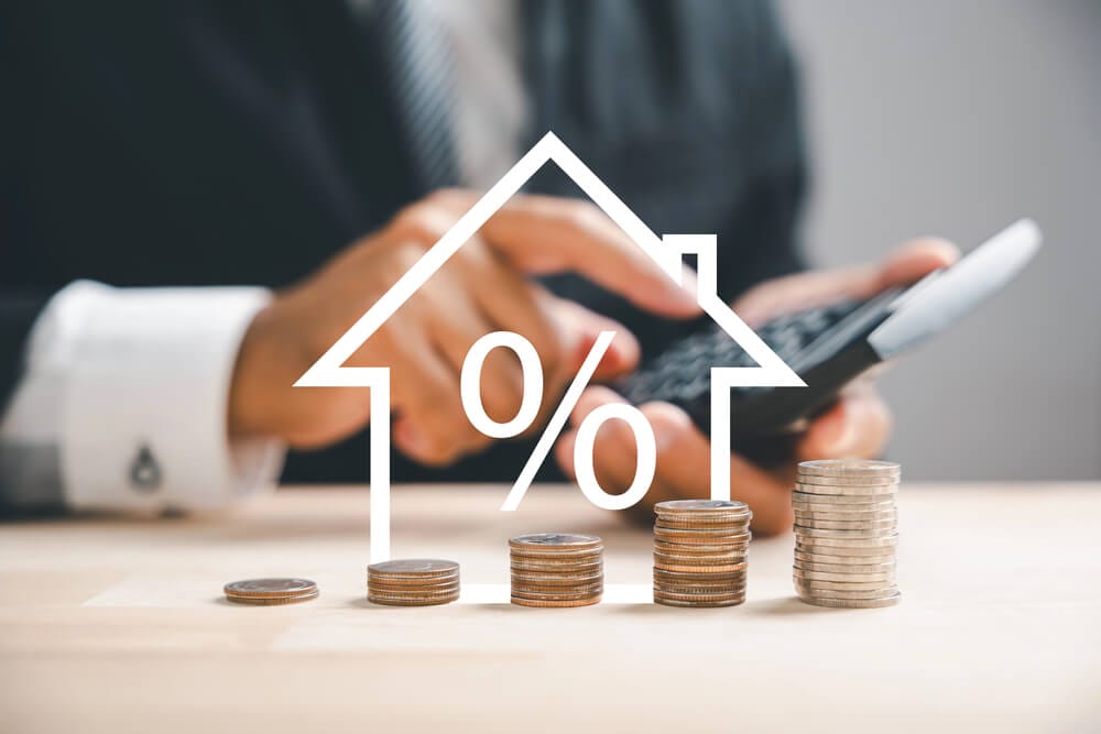 grafika przedstawiająca zarys domu, obok monety, w tle dłonie trzymające kalkulator